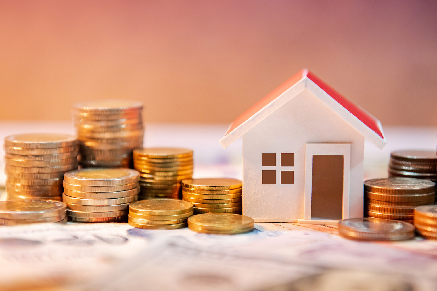 Symbolisches Hausmodell zwischen Münzstapeln auf Geldscheinen, Fokus auf Baufinanzierung oder Investitionen
