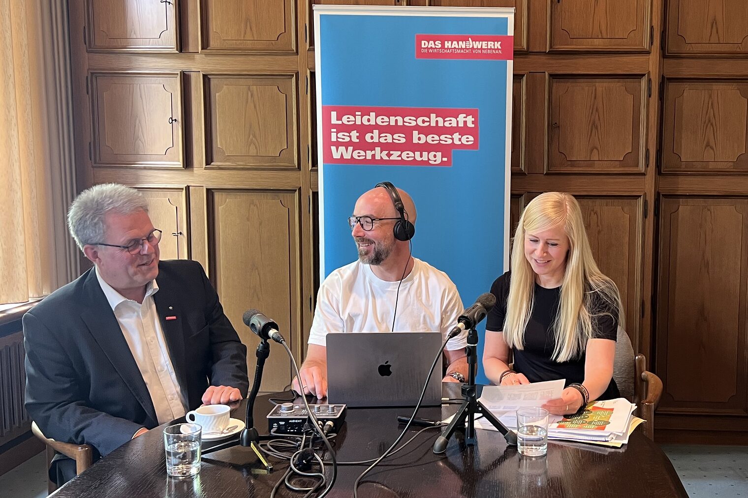 Drei Personen bei einer Podcast-Aufzeichnung am Tisch mit Mikrofonen, Laptop Roll-Up im Hintergrund.