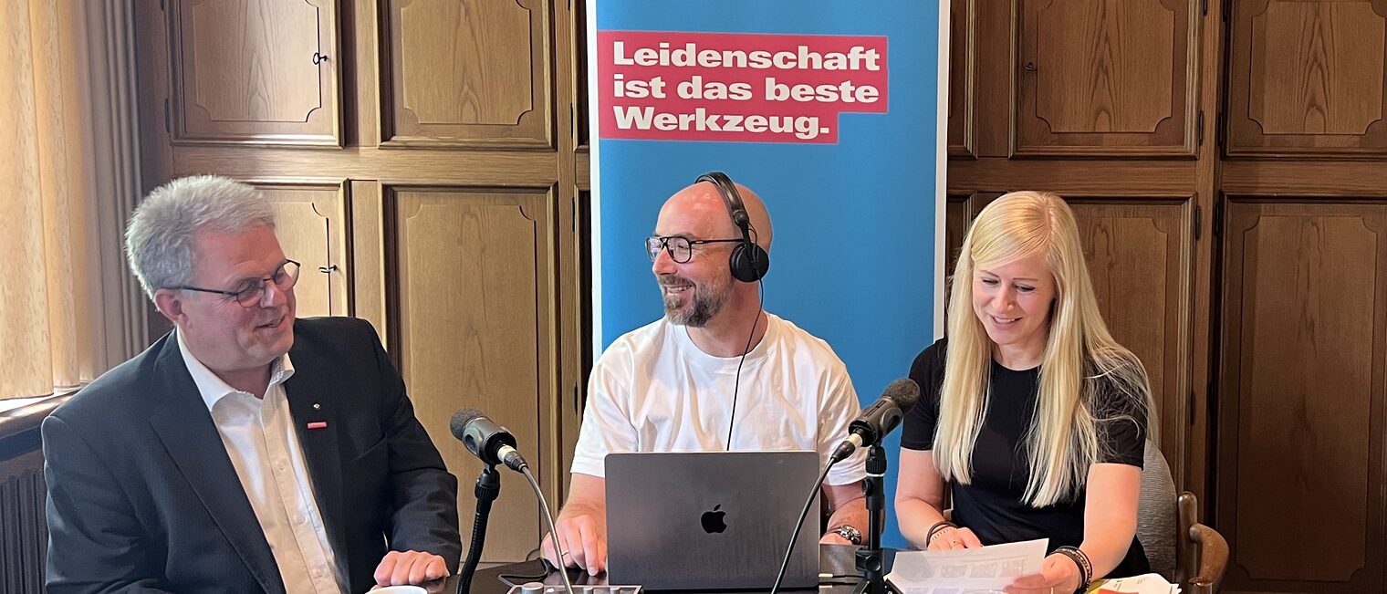 Podcast-Produktion mit Gästen aus dem Handwerk im Interviewformat Drei Personen bei einer Podcast-Aufzeichnung am Tisch mit Mikrofonen, Laptop Roll-Up im Hintergrund.